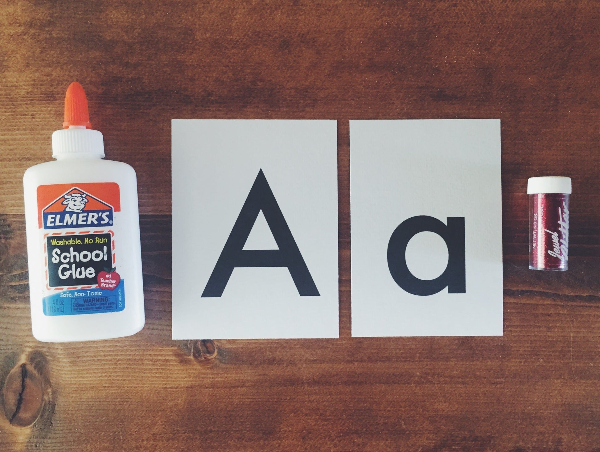 Letter 'A' cards next to red glitter and elmers glue on a wooden table.